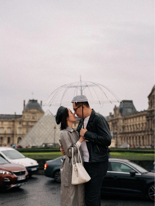 Couple sharing intimate moment under clear umbrella at the Louvre Museum Paris, rainy day engagement photo