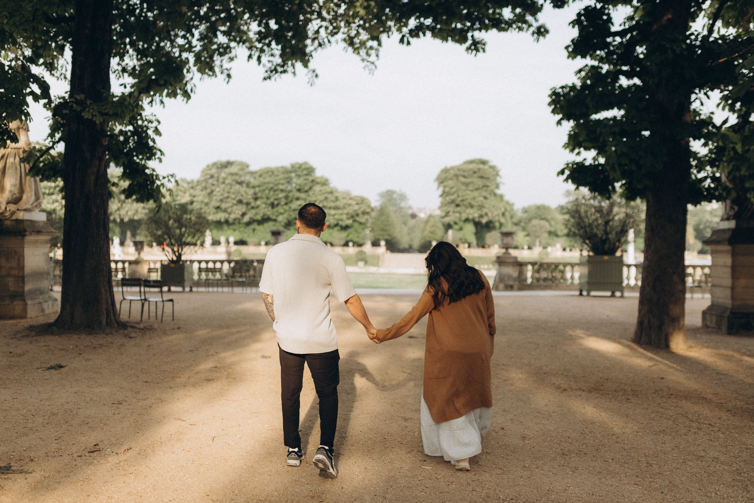 Couple walking hand in hand along a tree-lined path in the Luxembourg Gardens, seen from behind