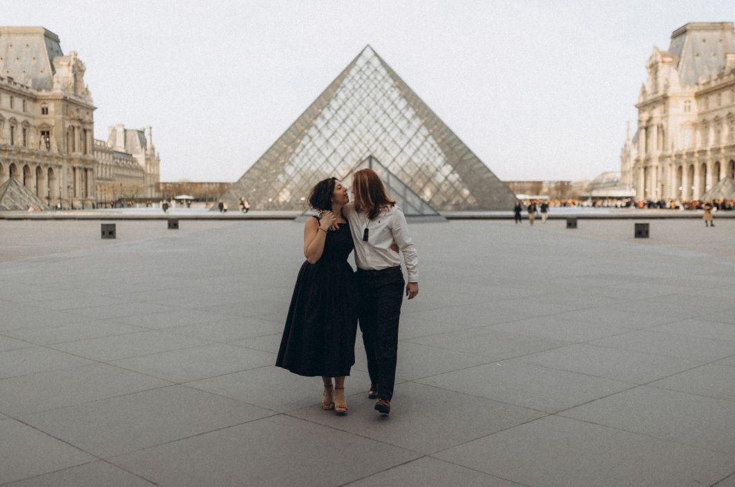Two women walking arm in arm in the Cour Napoléon with the Louvre Pyramid centered behind them, laughing together