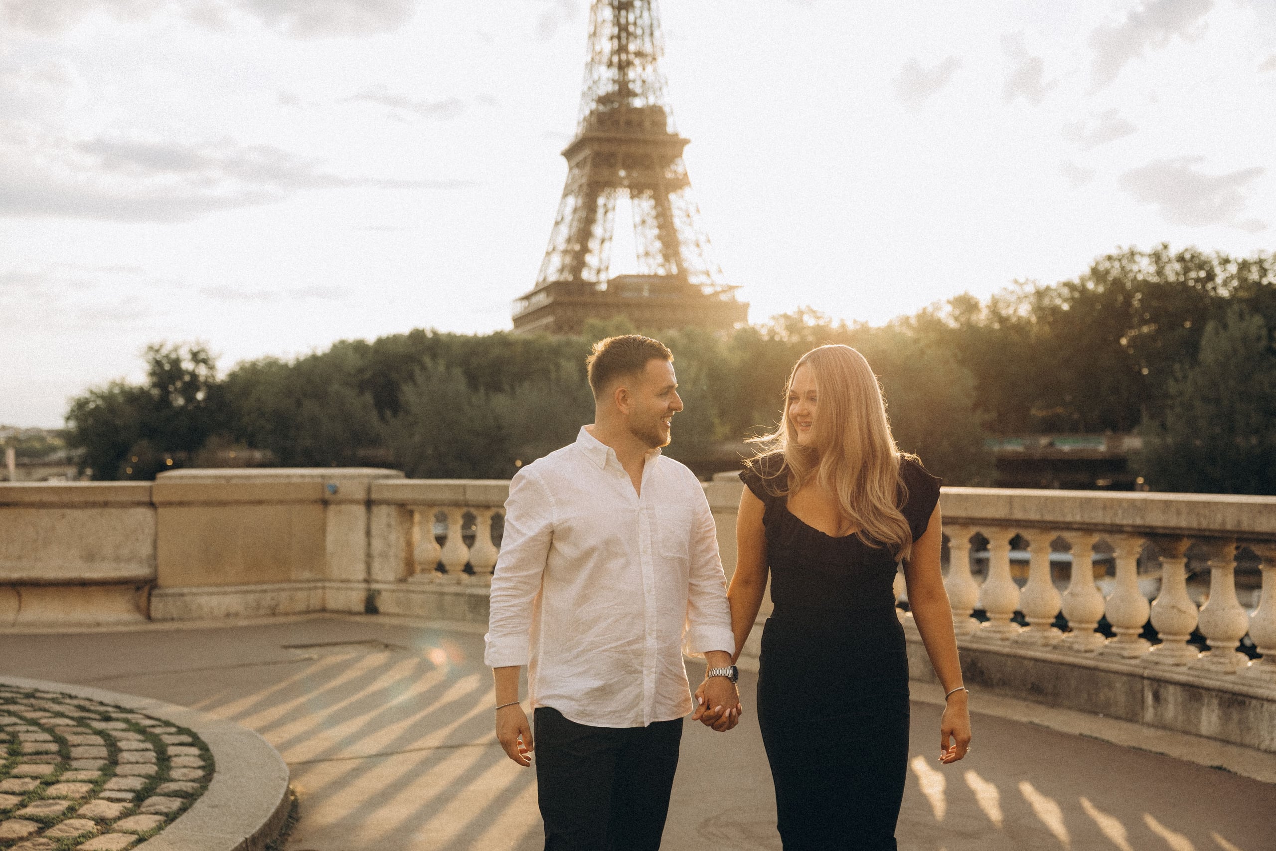 Couple walking hand in hand at golden hour with the Eiffel Tower in the background, smiling at each other