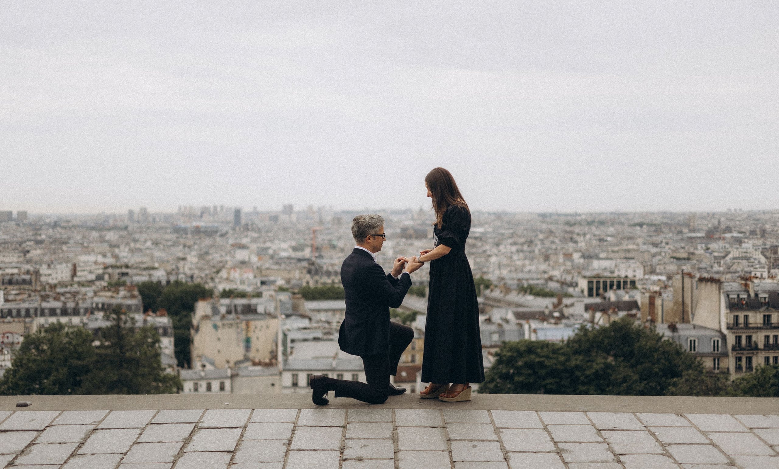 Man proposing on one knee to a woman at a Montmartre viewpoint with the Paris skyline in the background