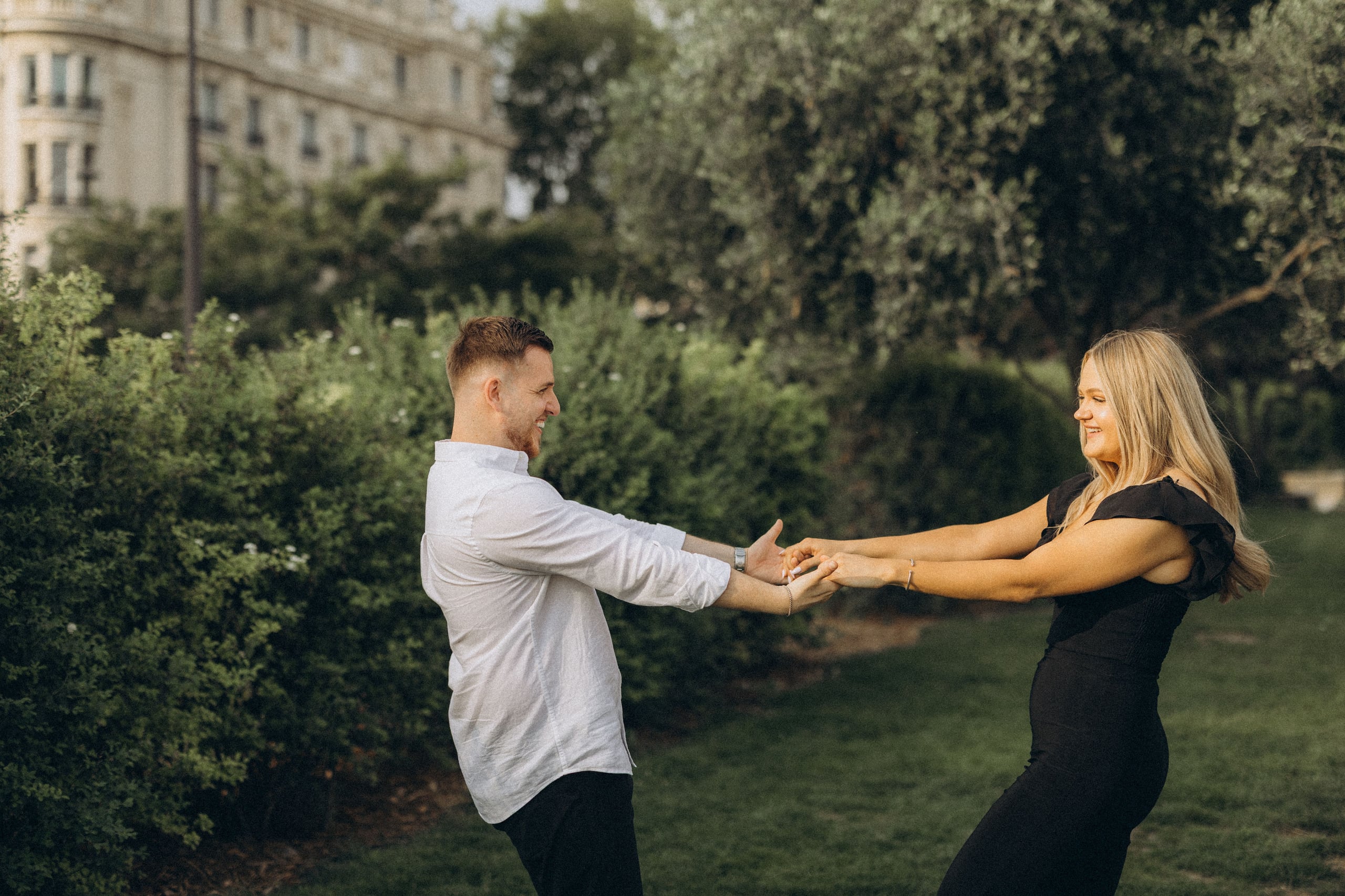 Couple playfully holding hands and leaning back in a green garden near Place des Vosges, laughing together